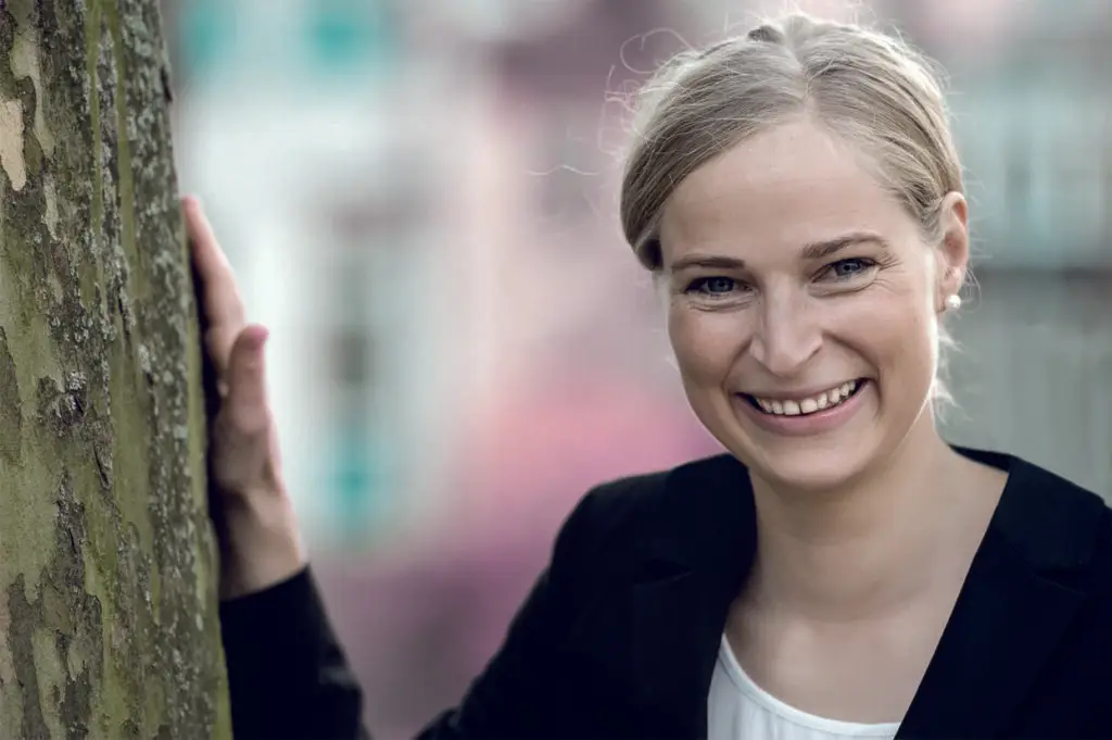 Frau lächelt im Freien an Baum gelehnt.